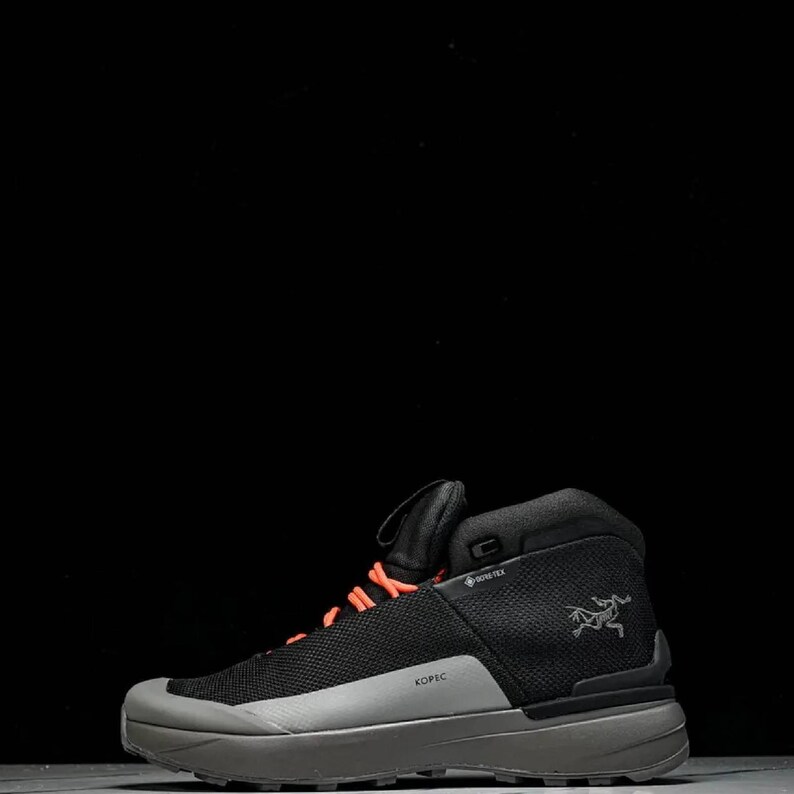 May include: Black and grey hiking boot with orange laces. The boot has a mesh upper, a grey sole, and the word "KOPEC" printed on the side. The Arc'teryx logo is visible on the side of the boot.