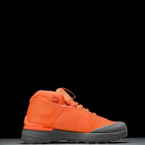 May include: An orange high-top trainer with a grey sole. The shoe has a mesh upper and a lace-up design. The trainer is set against a black background.
