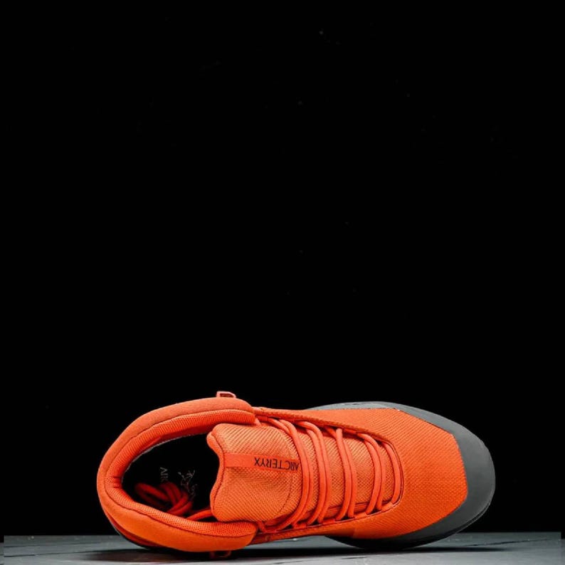 May include: Vibrant orange hiking boot with grey detailing. The boot has a mesh upper, a lace-up design, and the brand name "Arc'teryx". The sole is dark grey, and the boot is set against a black backdrop. A stylish and practical outdoor footwear option.