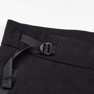 May include: Close-up of black pants with a black buckle and strap detail. The pants are made of a dark fabric and have a minimalist design. The buckle is centered on the waistband.