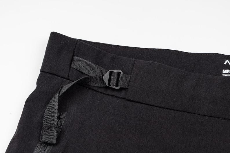May include: Close-up of black pants with a black buckle and strap detail. The pants are made of a dark fabric and have a minimalist design. The buckle is centered on the waistband.