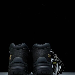 May include: Two black hiking boots with a textured upper and chunky soles. Each boot features a gold logo on the heel and a Gore-Tex tag. The background is a plain black.
