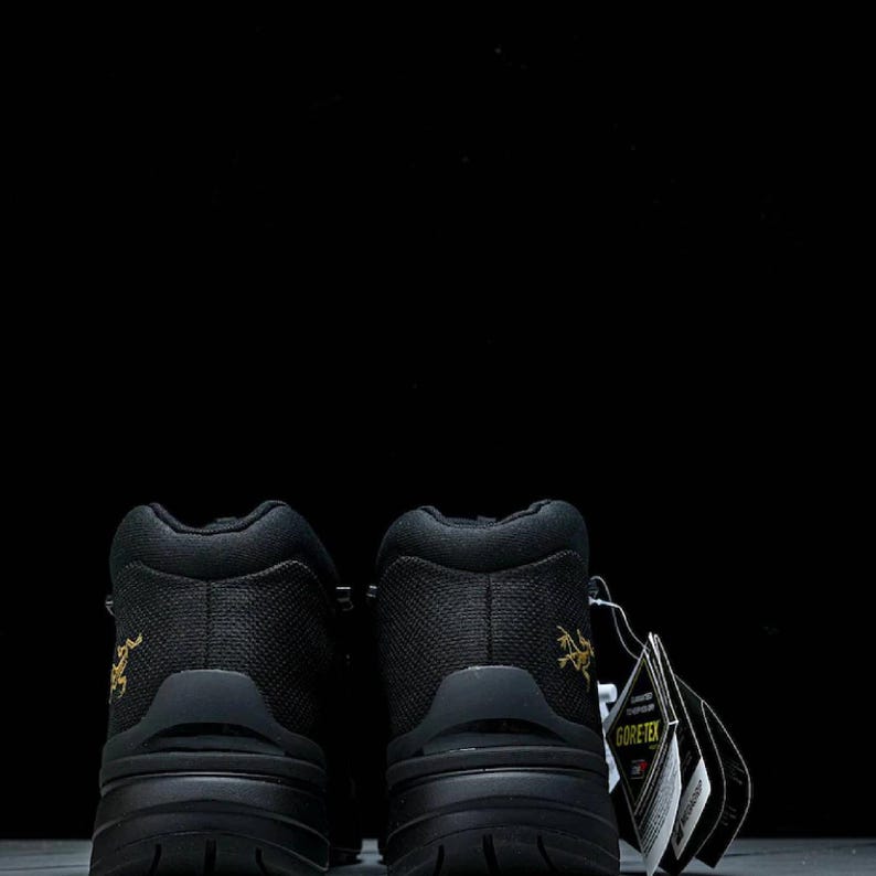 May include: Two black hiking boots with a textured upper and chunky soles. Each boot features a gold logo on the heel and a Gore-Tex tag. The background is a plain black.