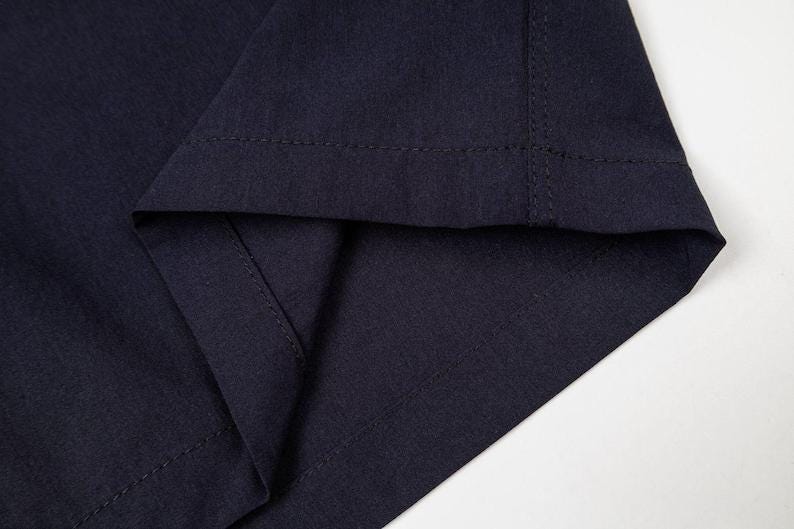 May include: Close-up of navy blue fabric, showing the folded hem and stitched seam. The fabric appears to be a medium-weight material, with a slight sheen. The image is well-lit, highlighting the texture and color of the textile.