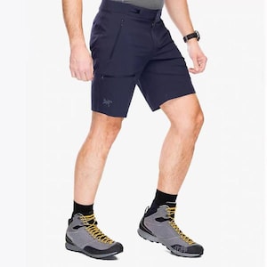 May include: Navy blue shorts with zippered pockets and a small logo on the left leg. The shorts are paired with gray and black hiking boots with yellow laces and black socks. A black watch is visible on the wrist.