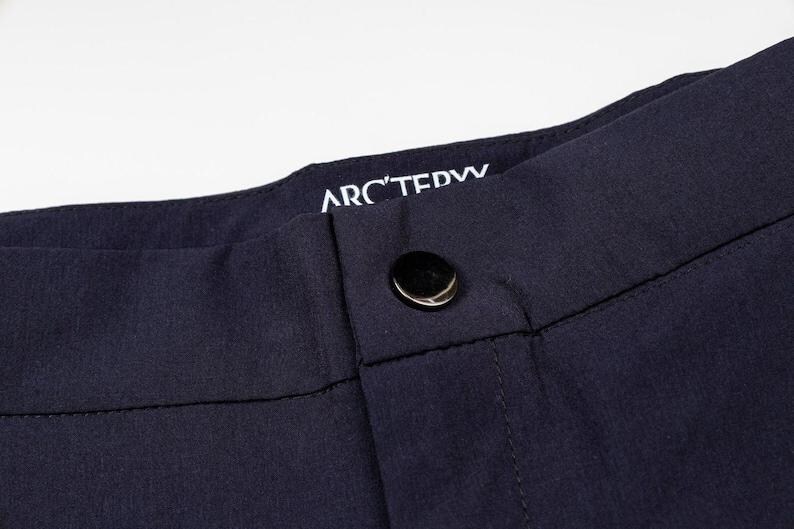 May include: Close-up of navy blue pants with a button closure. The brand name "ARC'TERYX" is visible in white lettering on the waistband. The pants appear to be made of a smooth, durable fabric.
