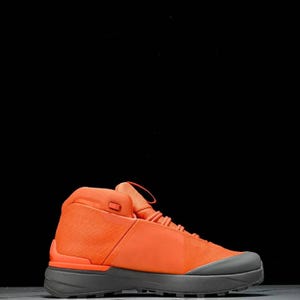May include: Orange high-top trainer with a grey sole. The shoe has a mesh upper, a padded collar, and a lace-up fastening. The design is modern and minimalist, suitable for various activities.