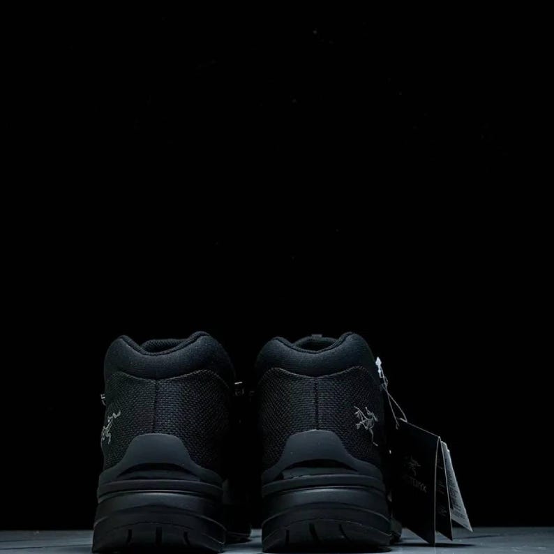 May include: Two black hiking boots with a textured upper and thick soles against a black backdrop. The boots have a high-top design and a small logo on the side. A tag is attached to one of the boots.