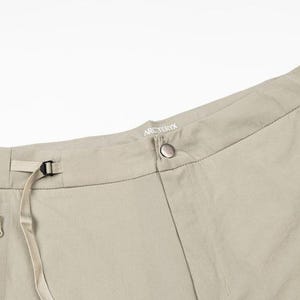 May include: Close-up of tan-colored outdoor pants. The pants have a button closure, a belt loop with a buckle, and two zippered pockets. The brand name "Arc'teryx" is visible on the waistband.