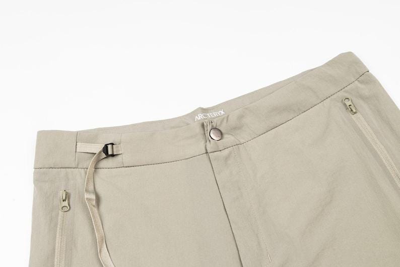 May include: Close-up of tan-colored outdoor pants. The pants have a button closure, a belt loop with a buckle, and two zippered pockets. The brand name "Arc'teryx" is visible on the waistband.