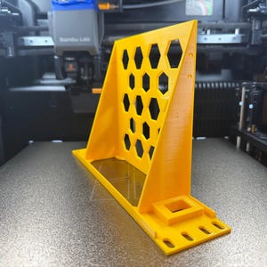 Könnte beinhalten: Ein leuchtend gelbes 3D-gedrucktes Objekt mit einem Wabenmuster und einer quadratischen Basis. Das Objekt befindet sich auf einer grauen Oberfläche, wahrscheinlich in einem 3D-Drucker. Das Design umfasst eine Reihe sechseckiger Aussparungen.