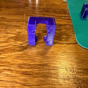Puede incluir: Un soporte azul vibrante impreso en 3D con un diseño en forma de U. El soporte tiene una forma rectangular con una abertura central y varios agujeros pequeños. El objeto está colocado sobre una superficie de madera, con una alfombra verde al fondo.