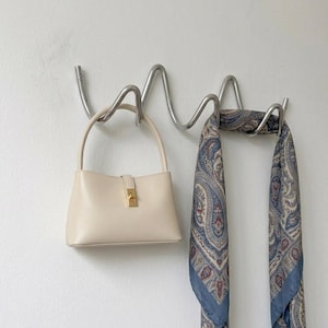 Puede incluir: Un bolso de color crema con una sola asa y un cierre rectangular en tono dorado, colgado de un gancho de pared de metal plateado ondulado junto a un pañuelo largo con un estampado de cachemira azul, gris y rojo sobre fondo claro.