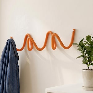 Könnte beinhalten: Eine leuchtend orangefarbene Wandgarderobe mit einem durchgehenden, fließenden, schlangenförmigen Design mit mehreren Schlaufen zum Aufhängen von Gegenständen. Die Garderobe ist mit kleinen runden Beschlägen an der Wand befestigt und hält eine blaue Jeansjacke.