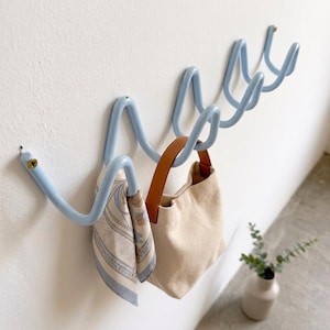 Könnte beinhalten: Eine hellblaue, wellenförmige Wandgarderobe aus Metall mit mehreren Haken. Ein gemusterter Seidenschal und eine kleine beige Segeltuchtasche mit braunen Ledergriffen hängen an den Haken. Die Garderobe ist mit sichtbaren Schrauben an der Wand befestigt.