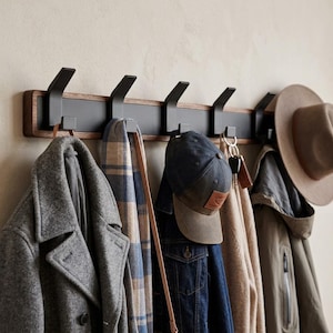 May include: A wall-mounted coat rack featuring a horizontal wooden base with a black centre panel and five matte black metal hooks. The rack holds a grey wool coat, a blue and beige plaid scarf, a denim jacket, a baseball cap, and a tan hat.