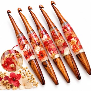 May include: Five crochet hooks with dark brown wood tips and handles, featuring a clear resin center section filled with embedded red, pink, and white dried hydrangea flowers. A circular inset shows a magnified view of the floral detail.