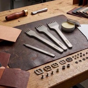 May include: A set of three silver-toned metal leather stitching chisels with 4mm markings, a thin metal awl, a wooden-handled mallet, a small round tin of wax, various metal buckles, rivets, and leather swatches on a dark brown leather mat.