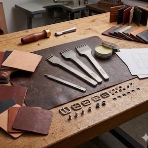 May include: A collection of leather crafting tools including three metal stitching chisels marked 4mm, a metal awl, a wooden burnisher, a tin of wax, various brass buckles, rivets, leather swatches, and finished leather wallets on a wooden table.