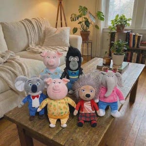May include: A group of plush character dolls from the animated film "Sing" are displayed on a wooden table. The dolls feature a koala, a gorilla, a pig, an elephant, and a lion, each with distinct outfits and expressions. The dolls are in various colours.