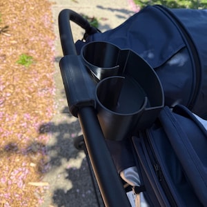 May include: A black stroller cup holder with two circular compartments and a curved design. The cup holder is attached to the stroller's handle. The stroller is navy blue, and the background shows a pavement and fallen pink flower petals.