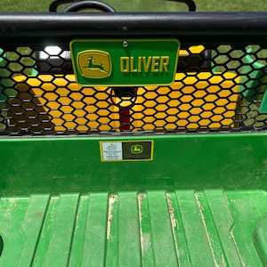 Puede incluir: Una caja de carga de vehículo utilitario verde y amarilla con una rejilla negra de panal. Un letrero rectangular verde con el logotipo amarillo de John Deere y el nombre OLIVER en letras amarillas está fijado al centro de la rejilla.