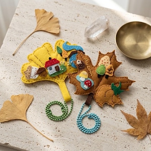 May include: Two felt ornaments shaped like leaves. One is a yellow ginkgo leaf with a small house, acorns, and a blue stream. The other is a brown maple leaf with the word Autumn, mushrooms, and a pumpkin. Both have green beaded chains.
