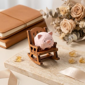 Pode incluir: Uma pequena estatueta de porco em madeira esculpida em rosa claro com uma superfície lisa e facetada e olhos pretos simples, sentada numa cadeira de balanço em miniatura de madeira castanha. A cadeira apresenta um encosto de ripas.