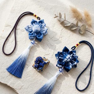 May include: Two decorative fabric flower ornaments featuring blue and white tie-dye patterns, each attached to a dark blue cord loop. The flowers are accented with clear beads and long, ombre-style blue and white tassels that fade in color.