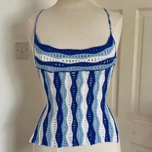 May include: A blue, white, and light blue crochet top with thin straps. The top features a wavy pattern in shades of blue and white. The top is displayed on a mannequin.