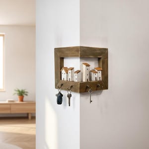 Peut inclure: Un porte-clés d'angle en bois avec une finition marron rustique et une rangée décorative de cinq maisons miniatures blanches aux toits marron. L'unité comprend quatre crochets en métal sur le bord inférieur, retenant actuellement trois clés.