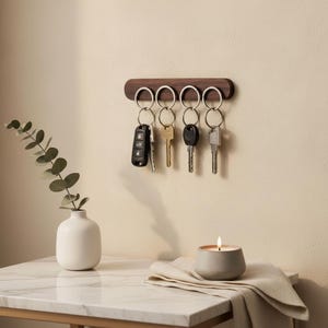 Peut inclure: Un porte-clés mural horizontal en bois brun foncé doté de quatre anneaux métalliques argentés. Chaque anneau retient un trousseau de clés, incluant une télécommande de voiture noire, une clé en laiton et deux clés métalliques argentées.