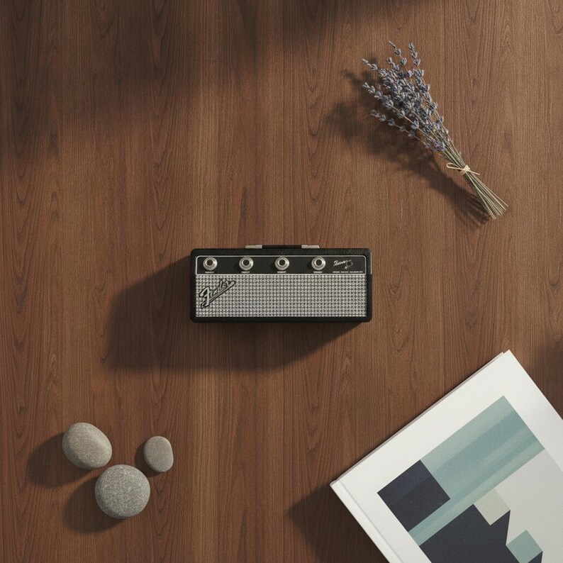 May include: A black Fender guitar amplifier with a silver mesh grille, four control knobs, and the Fender logo on the front. The amplifier is positioned on a wood grain surface alongside a small bundle of dried lavender, three smooth gray stones, and a book.