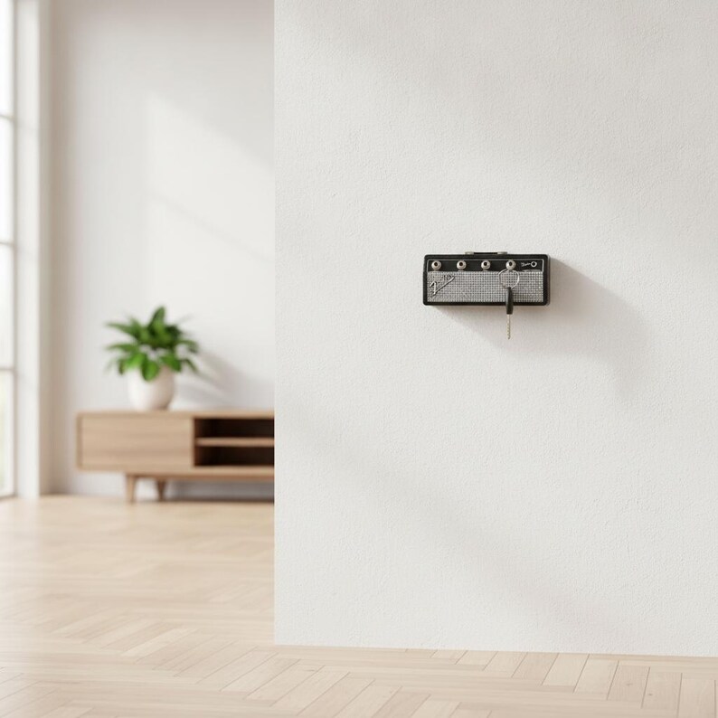 May include: A black wall-mounted key holder designed to resemble a miniature guitar amplifier. The unit features a silver speaker grille, four control knobs along the top edge, and a Fender logo on the left side, with a single metal key inserted.