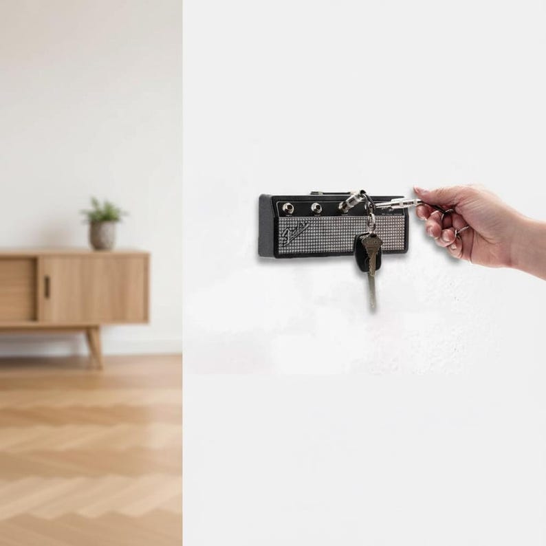 May include: A black and silver wall-mounted key holder designed to resemble a miniature guitar amplifier. The unit features a textured silver grille, black casing, and the Fender logo. It includes input jacks for inserting keychains.
