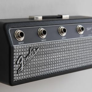 May include: A black Fender guitar amplifier wall mount with a silver woven grille cloth front. The unit features four circular input jacks along the top control panel, each labeled with the word INPUT, and a black carrying handle on the top edge.