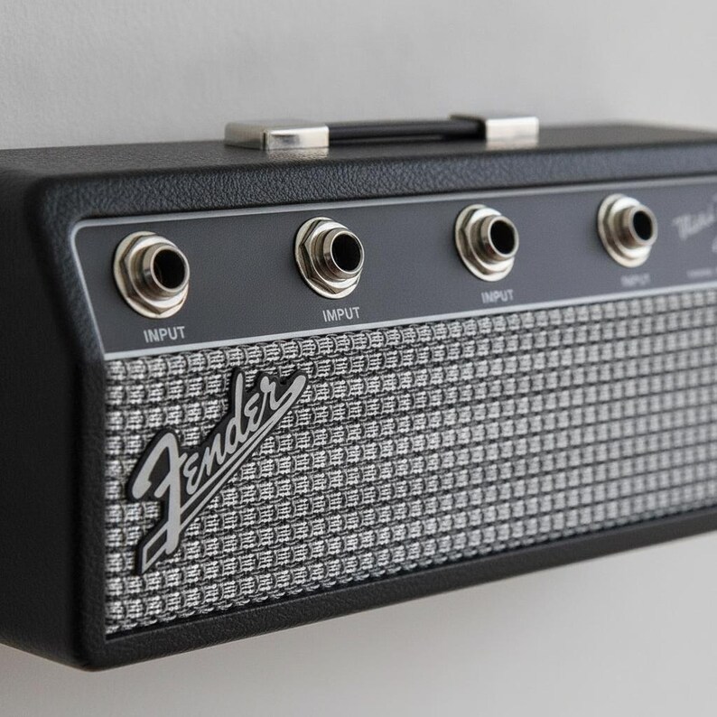 May include: A black Fender guitar amplifier wall mount with a silver woven grille cloth front. The unit features four circular input jacks along the top control panel, each labeled with the word INPUT, and a black carrying handle on the top edge.