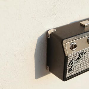 May include: A black, rectangular guitar amplifier key holder with a textured finish, silver metal corner protectors, and a silver control panel featuring an input jack. The front grille displays the Fender logo in a classic script font style.