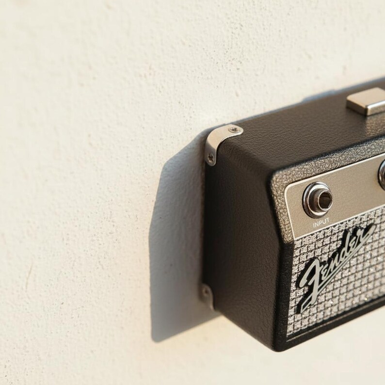 May include: A black, rectangular guitar amplifier key holder with a textured finish, silver metal corner protectors, and a silver control panel featuring an input jack. The front grille displays the Fender logo in a classic script font style.