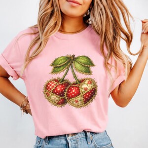 May include: A light pink short-sleeved t-shirt featuring a central graphic of two cherries. The cherries are designed with a patchwork aesthetic, incorporating various red and green patterns, polka dots, and floral prints with visible stitching.