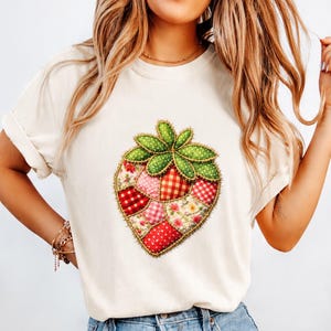 May include: A cream-coloured short-sleeve t-shirt featuring a large central graphic of a strawberry constructed from various patchwork fabric patterns including red gingham, floral prints, and polka dots, all outlined with a stitched effect.