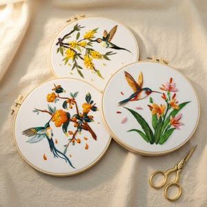 May include: Three circular wooden embroidery hoops containing white fabric with detailed needlework. The designs feature colourful hummingbirds interacting with yellow, orange, and pink floral branches. A pair of gold-toned metal scissors is included.