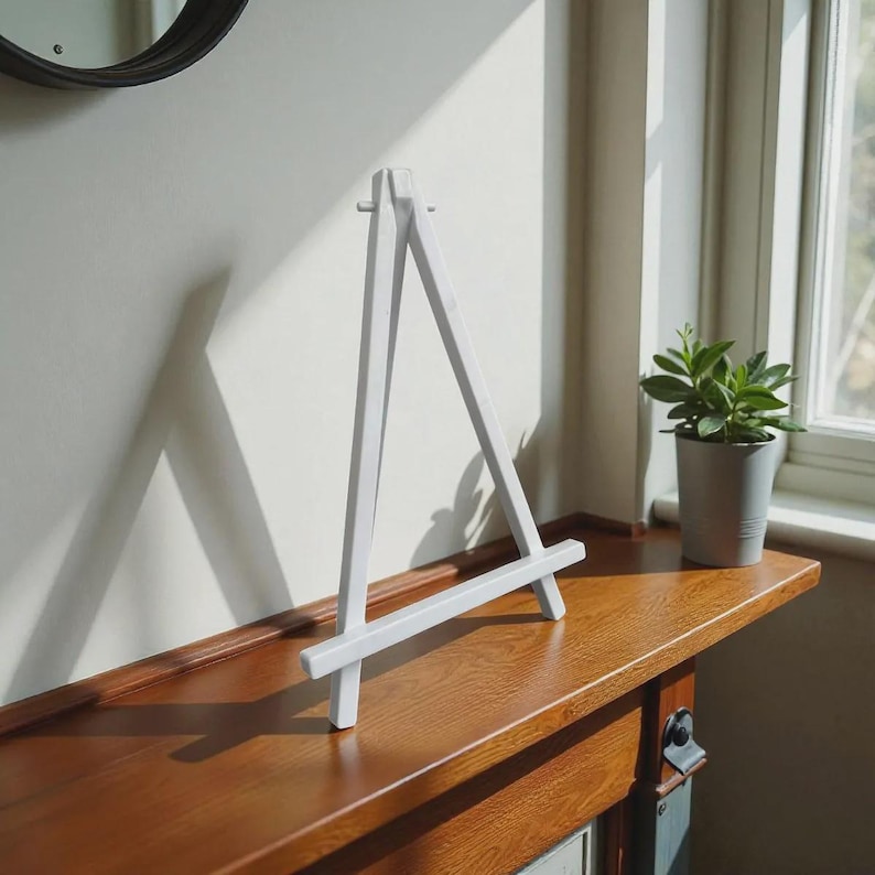 May include: A white wooden tabletop easel with a triangular frame design, featuring two long vertical legs connected by a horizontal support bar at the base and a small peg near the top to hold items securely in an upright display position.