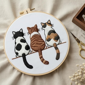Könnte beinhalten: Ein runder Stickrahmen aus Holz mit weißem Stoff, der ein handgesticktes Motiv von drei nebeneinander sitzenden Katzen zeigt. Die Katzen sind von hinten dargestellt, darunter eine schwarz-weiß gefleckte Katze, eine braune getigerte Katze und eine Glückskatze.