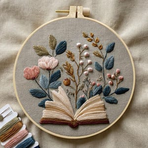 Puede incluir: Diseño floral bordado en un aro de madera. La obra presenta un libro abierto rodeado de flores y follaje en tonos rosa, azul, verde y marrón. Se ven hilos de bordar en colores coordinados.