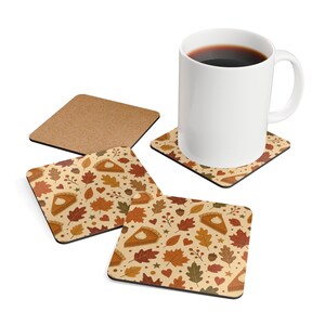 May include: A white ceramic mug and three square coasters featuring a repeating autumn pattern of orange pumpkin pie slices, brown and yellow fall leaves, small acorns, and red hearts on a light beige background. One coaster is flipped over.