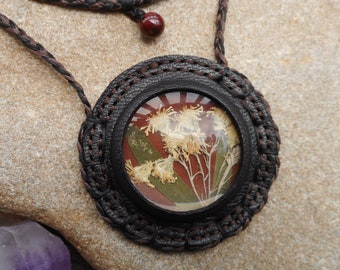 Op de afbeelding: Een bruine leren ketting met een ronde hanger met een geperst bloemenmotief. De hanger heeft een donkerbruin leren frame en een transparante glazen koepel. De bloemen zijn wit en geel met groene bladeren.