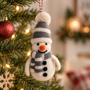 May include: A felt snowman ornament featuring a white body with three black buttons, a gray and white striped scarf, and a matching gray and white striped hat topped with a white pom-pom. The snowman has a small orange nose and a black stitched smile.
