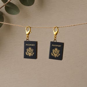 May include: Two miniature passport charms featuring dark blue covers with gold-colored text and the United States seal. Each charm is attached to a gold-toned lobster clasp, suspended from a thin string, showcasing detailed travel-themed accessories.