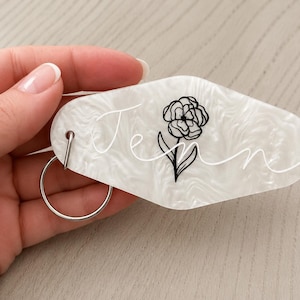 May include: A white pearlescent acrylic motel-style keychain with a diamond-like shape. It features the name Jenn written in white cursive script and a black line-art illustration of a flower centered on the surface with a silver metal ring.